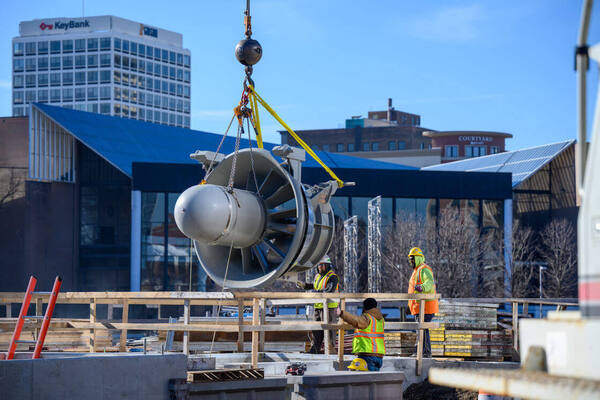Crane lowers a large gray turbine at the Seitz Park hydroelectric dam site. Three workers in high-vis vests guide it. KeyBank building visible.