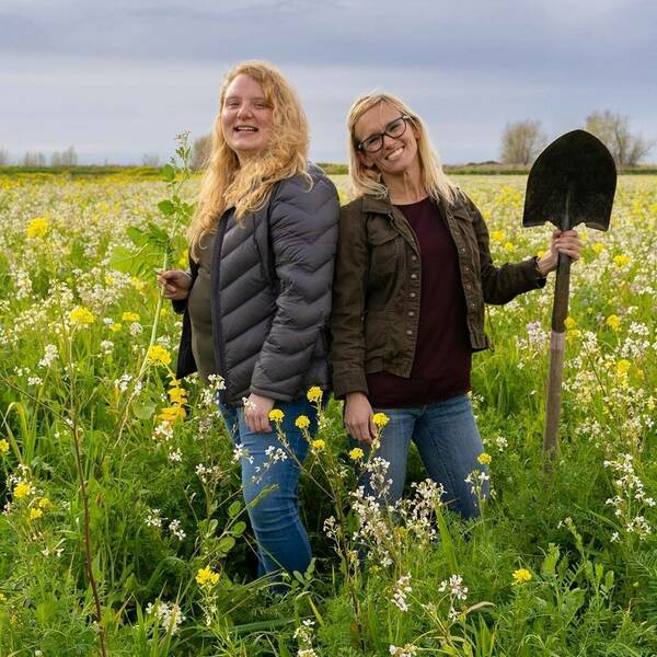 two caucasian women standing in a field of wildflowers looking at the camera and smiling with garden tools