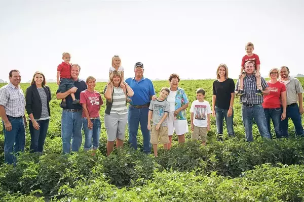 A large family group of men, women, and children stands in a field of tomato crops, some adults holding younger children on their shoulders.  They are casually dressed, and several are smiling.