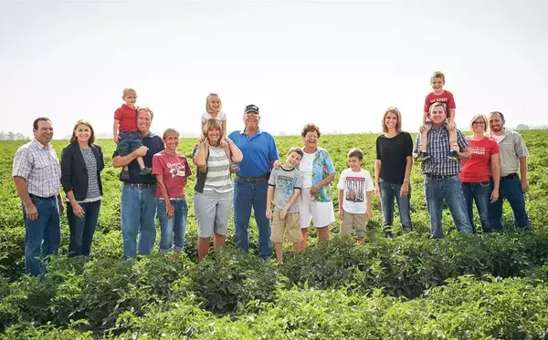 A large family group of men, women, and children stands in a field of tomato crops, some adults holding younger children on their shoulders. They are casually dressed, and several are smiling.