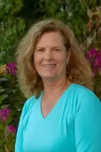 Woman with shoulder-length, wavy blonde hair, wearing a turquoise v-neck shirt, smiles in front of flowering bushes.