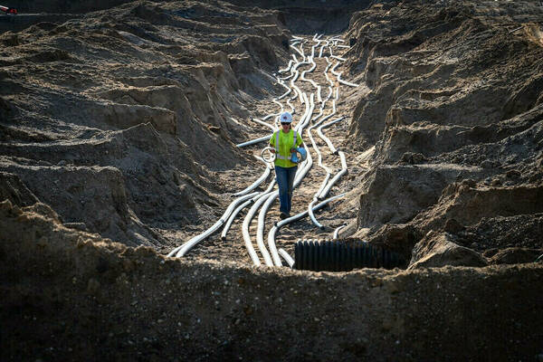 Construction worker in a neon yellow vest and white hard hat walks through a trench carrying conduit amidst numerous parallel lengths of grey conduit against dark brown dirt.