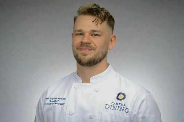 Jacob Eigenheer, Sous Chef at the University of Notre Dame, a young man with light brown hair and a beard, smiles while wearing a white chef's coat with the Campus Dining logo.