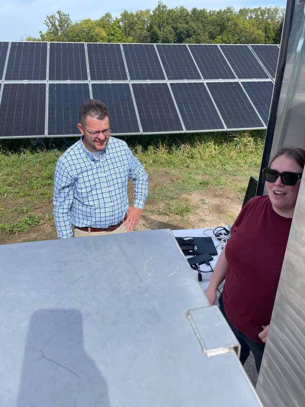 Man in blue plaid shirt smiles, woman in sunglasses and maroon shirt with open mouth. Solar panels in grassy area behind them.