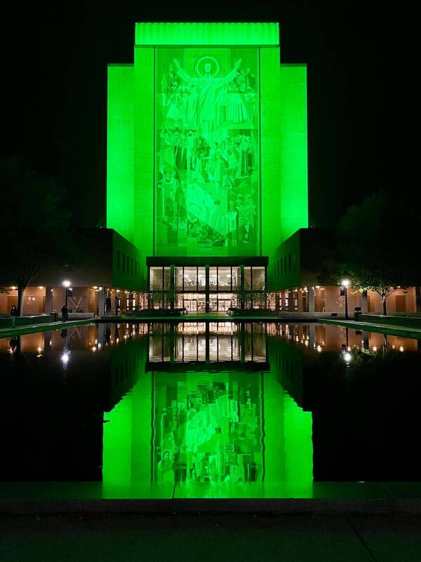 Hesburgh Library's "Word of Life" mural glows vibrant green at night, reflected perfectly in the reflecting pool below.