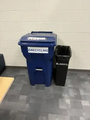 Blue "RECYCLING" bin and black "LANDFILL" bin against a white wall on a gray tiled floor.